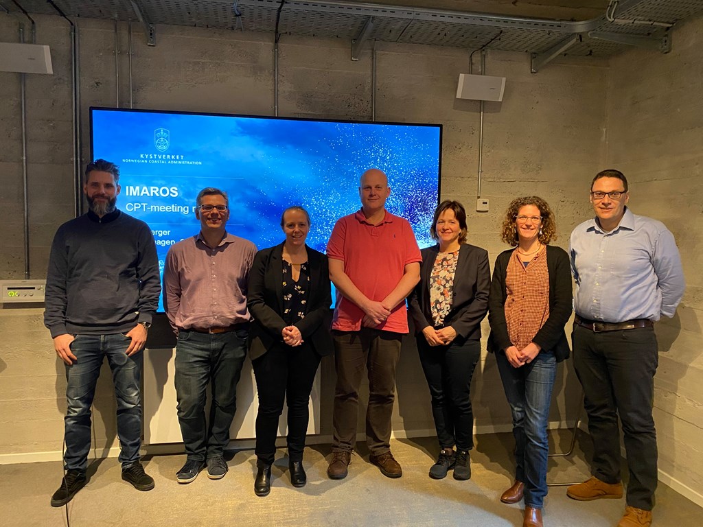 Syv mennesker som står på rad foran en skjerm. 