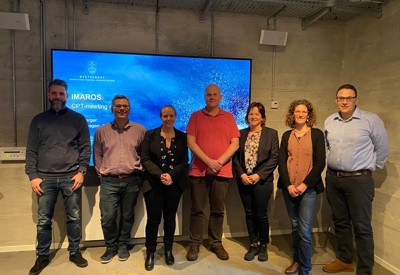 Syv mennesker som står på rad foran en skjerm.